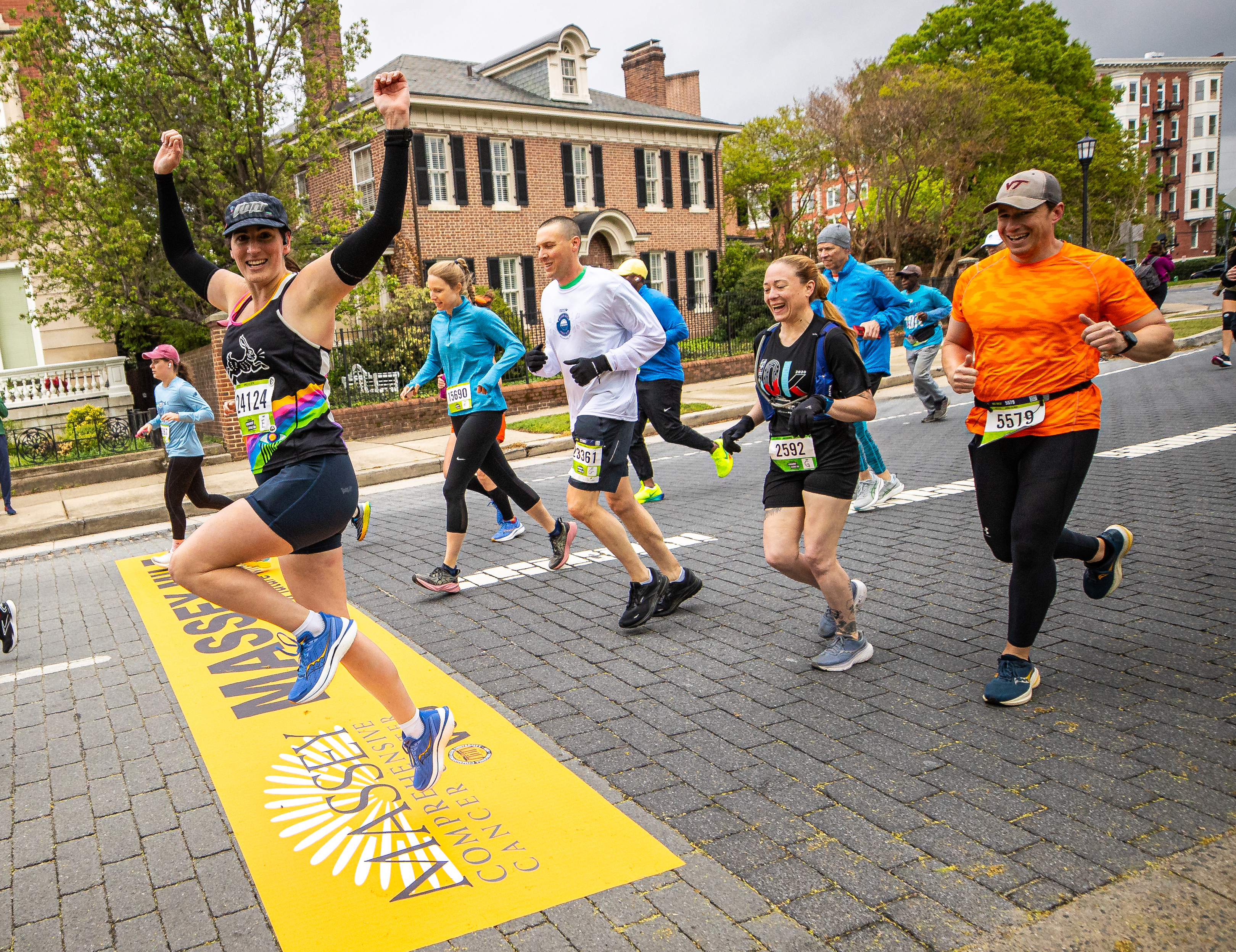 A group of diverse runners participating in a race on a cobblestone city street. In the foreground, a woman in a black tank top with a rainbow design cheers with her arms raised as she crosses a yellow 