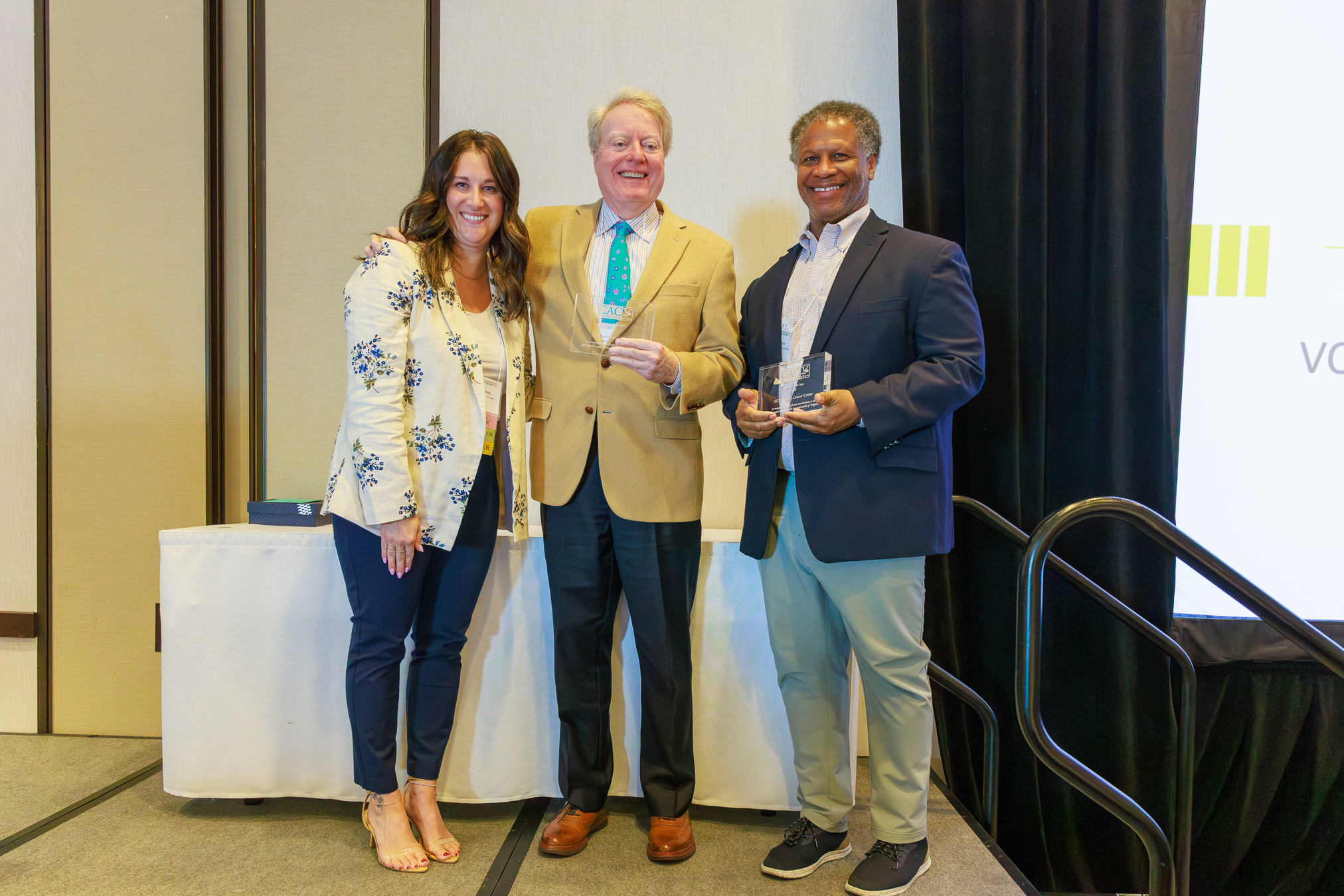 Robert Winn, Thomas Loughran, and Jessica Deering pictured on stage while Robert and Thomas hold rectangle, CACV-branded see-through awards