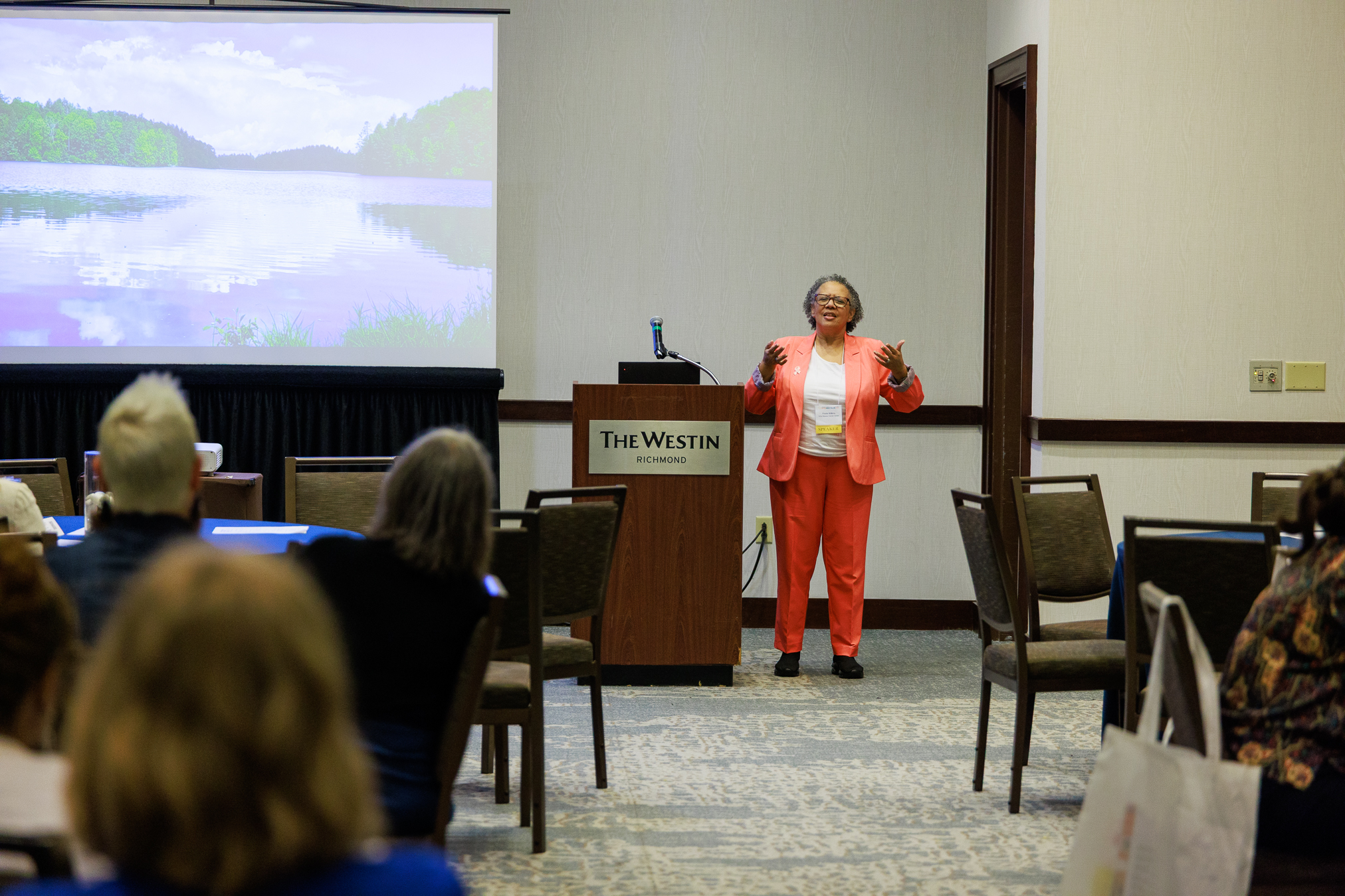 Freda Wilkins, M.S.W., standing in orange suit speaking at podium with hands slightly raised
