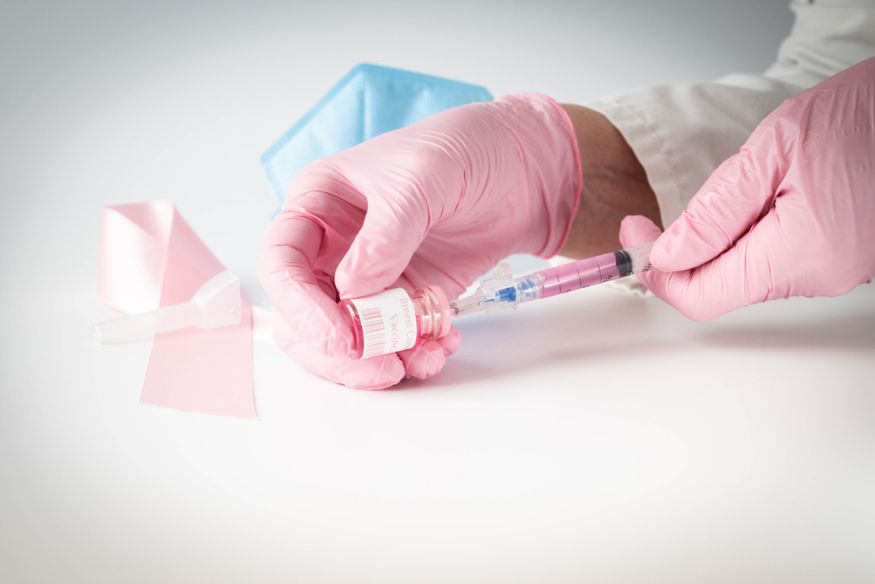 Pink gloved hands drawing up a dose of breast cancer vaccine from a glass vial, mock-up, pink vial.
