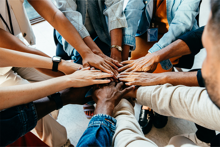 A diverse group of people put their hands together in the center of a circle, symbolizing teamwork, unity, and collaboration.