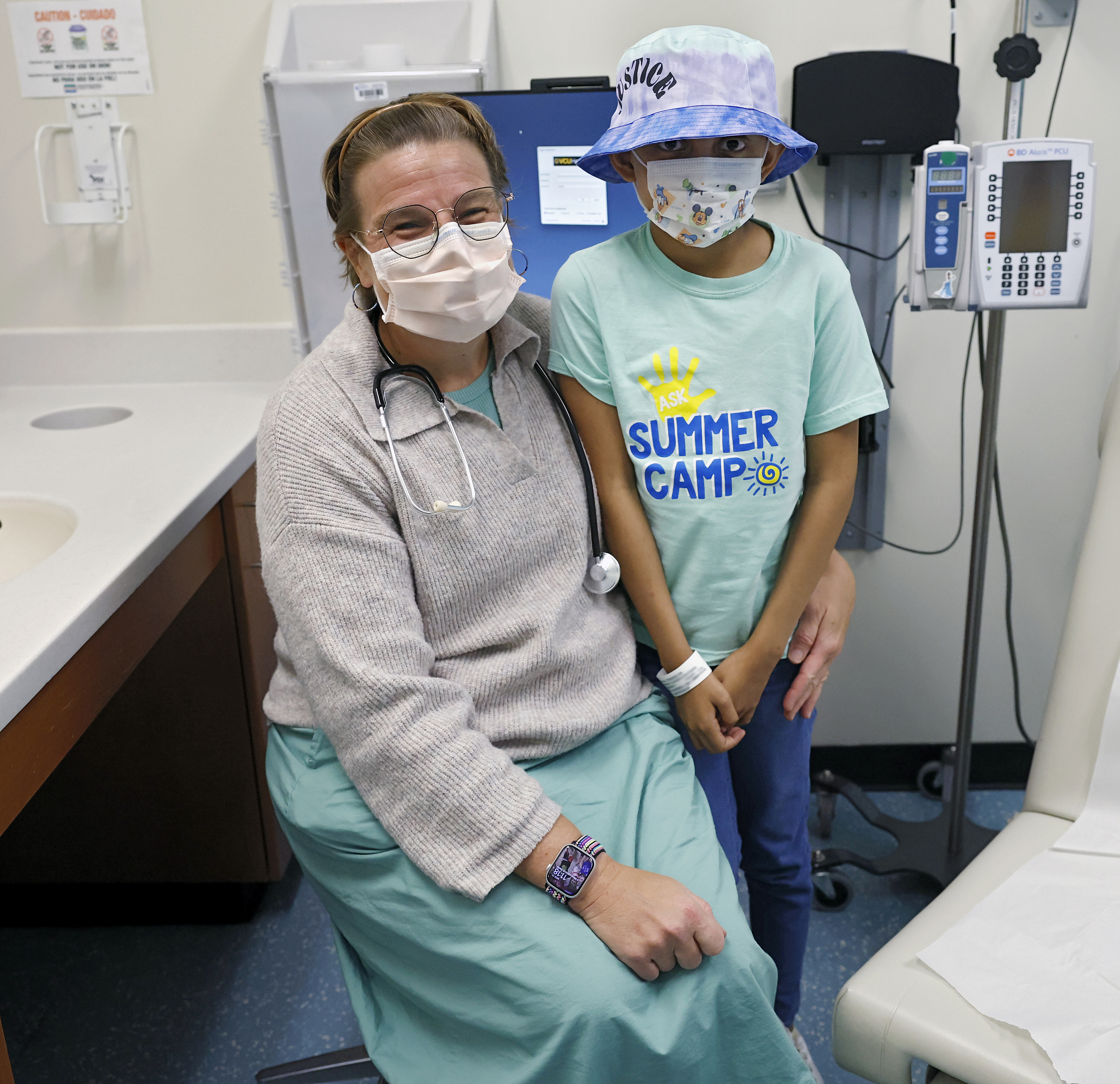 A doctor, Dr. Austin, and Estrella, a young pediatric patient, pose in an exam room. Estrella wears a bucket hat and a mask over a T-shirt that says 