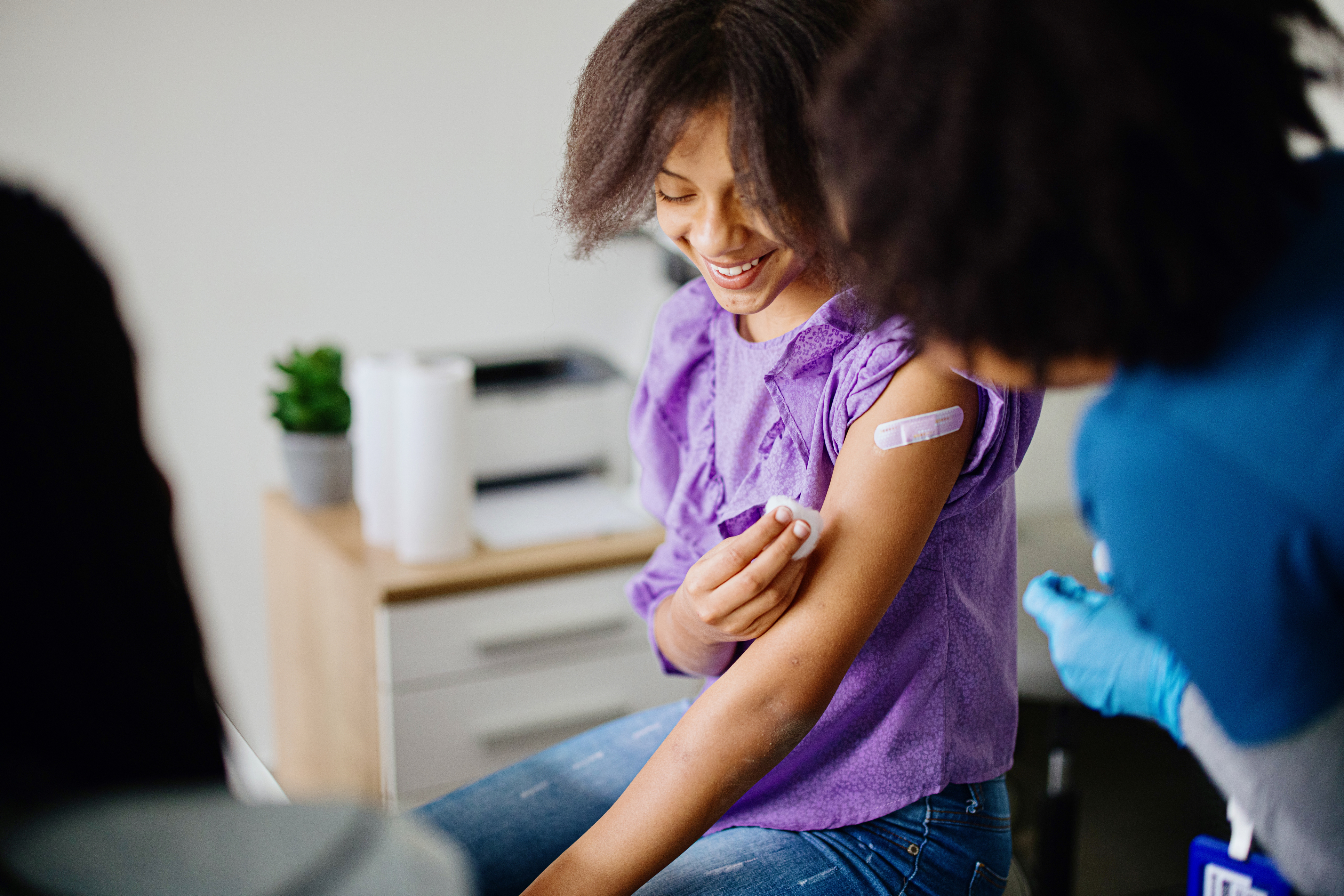 A smiling young girl in a purple shirt and jeans applies a cotton swab to a small bandage on her upper arm, likely after receiving the HPV vaccine, while a healthcare worker in blue scrubs and gloves looks on.