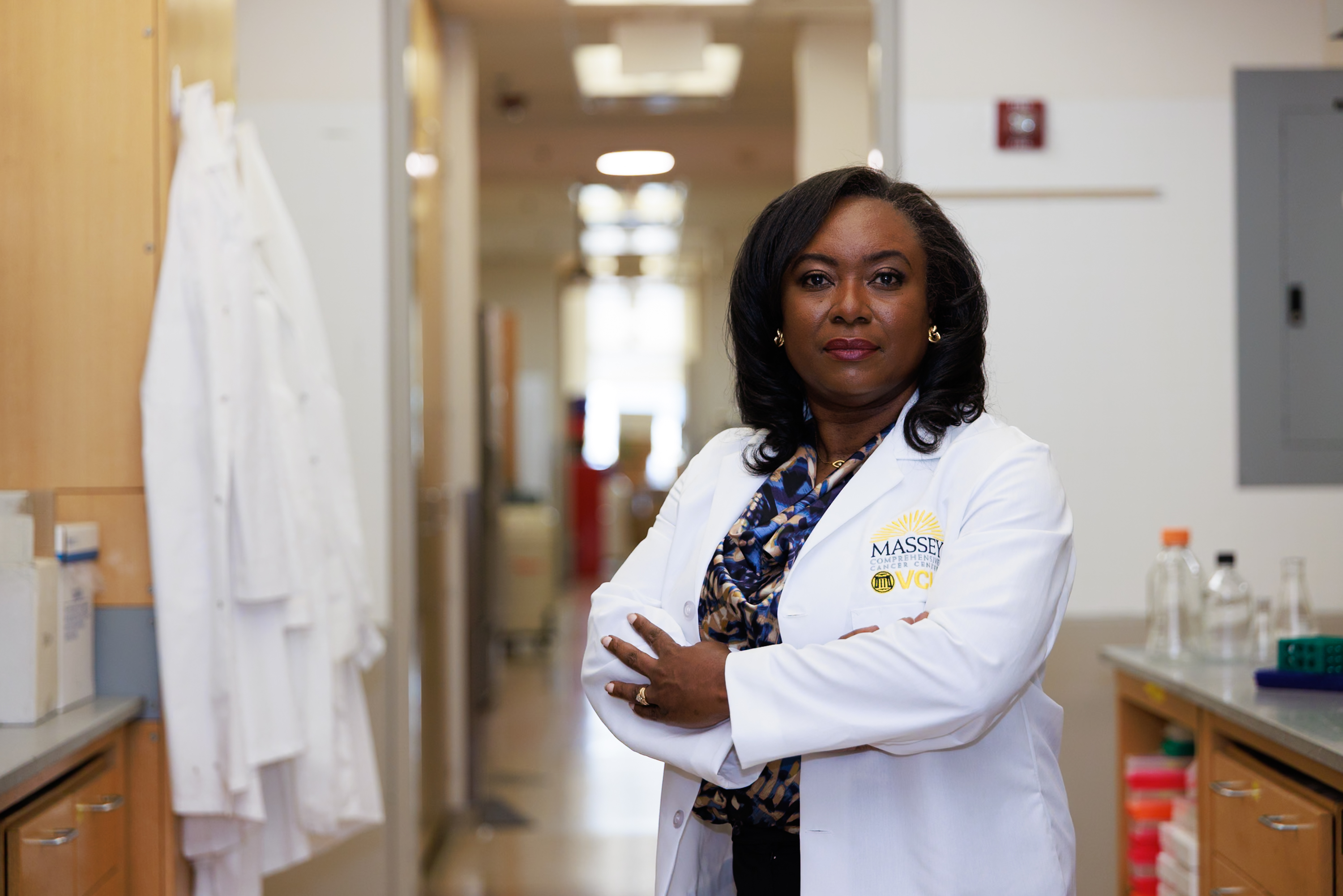 Portrait of Monica Baskin, Ph.D., standing with arms crossed in a laboratory corridor wearing a patterned blouse and a white lab coat with the VCU Massey Comprehensive Cancer Center logo.