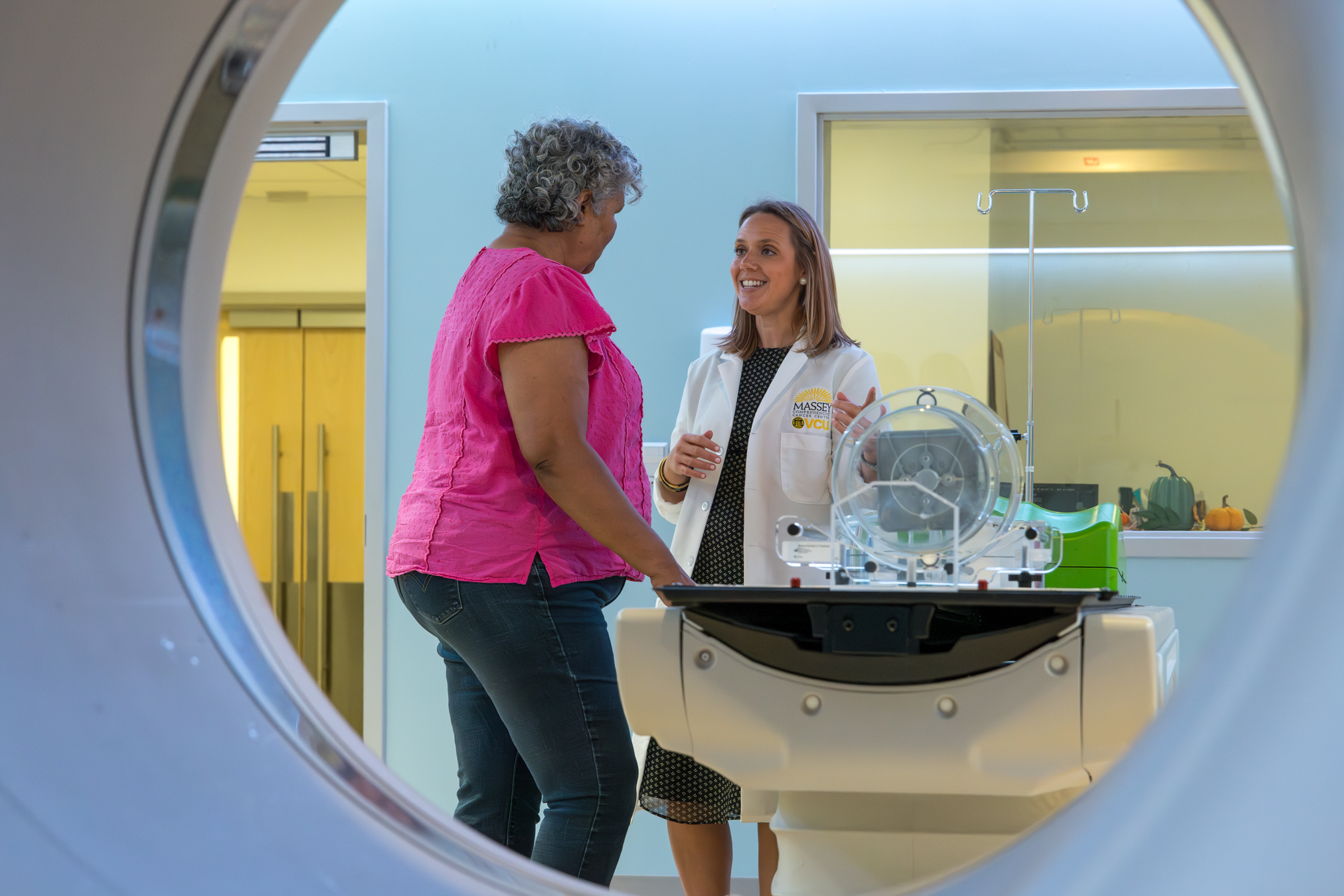 Bridget Quinn wearing a white lab coat and professional attire, smiles while speaking with a patient in a bright, modern medical imaging suite. They are standing next to a CT or PET scan machine, seen through the circular frame of the scanner's bore. In the background, a large window reveals a clean clinical area with an IV pole and small decorative pumpkins on a sill.
