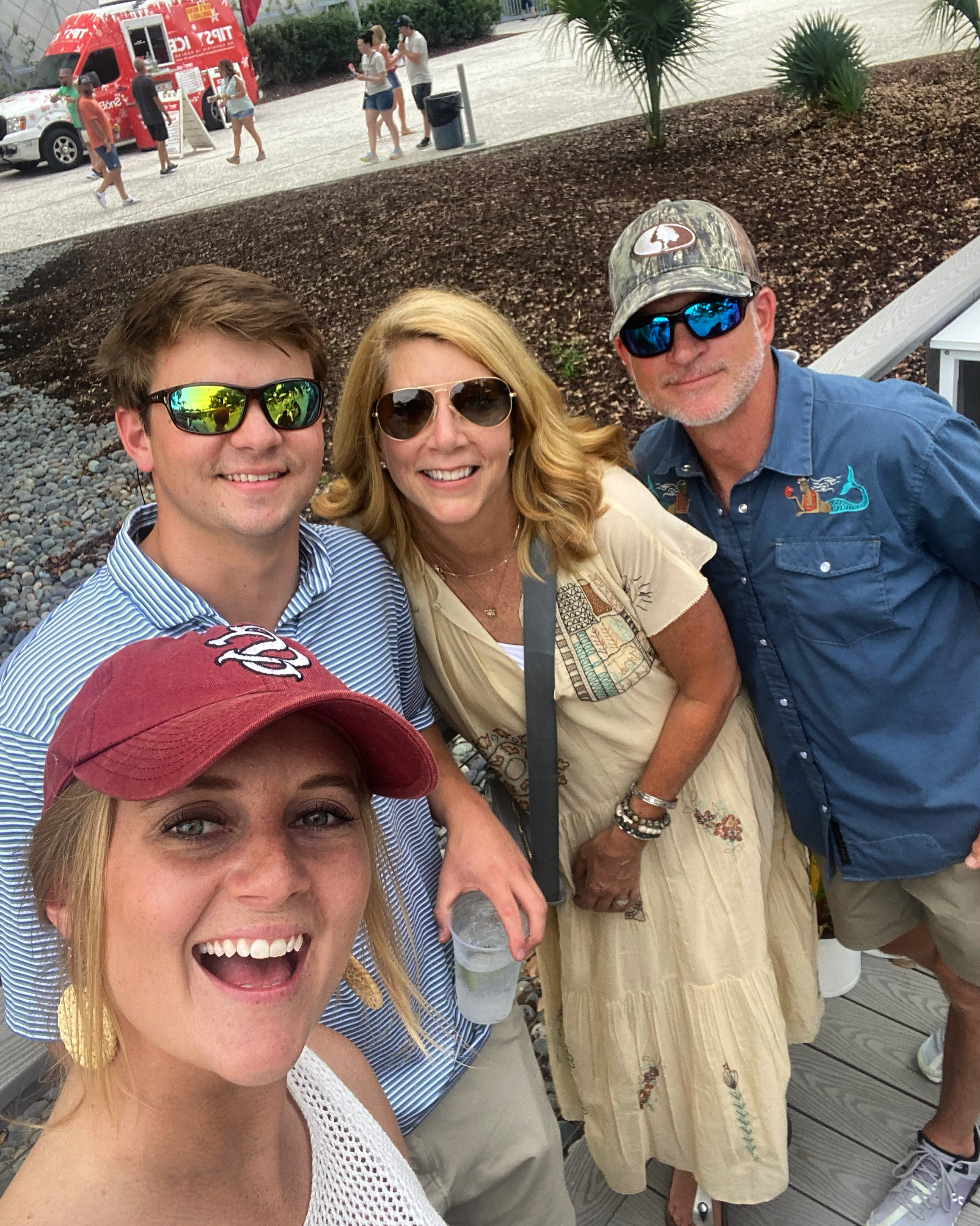 A group of four smiling people—two men and two women—posing for a selfie outdoors. In the background, there is a red food truck and people walking around an open plaza.