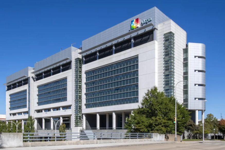 An exterior, eye-level shot of the modern Altria building under a clear blue sky. The multi-story structure features white facades, large grid windows, and silver panels, with the colorful Altria logo displayed prominently on the top right corner. A concrete overpass with a metal railing runs along the foreground, and a few green trees stand beside a street lamp on the right.
