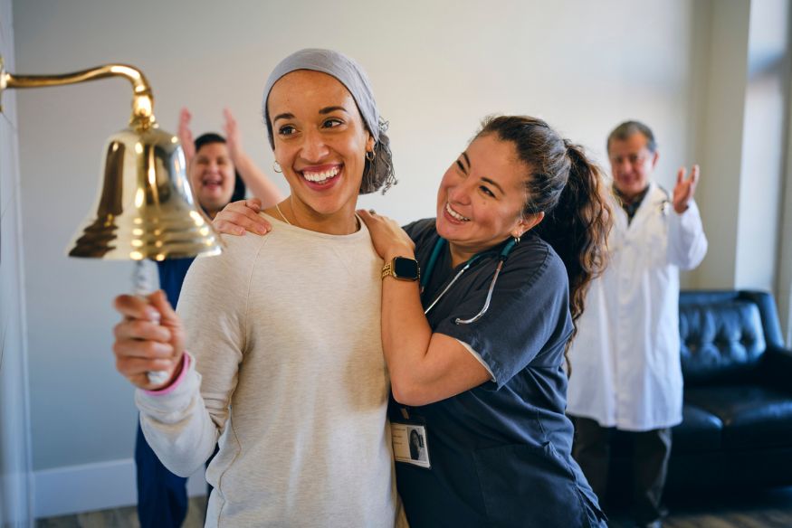 A smiling woman wearing a headscarf rings a brass bell on a wall while a nurse and medical staff applaud in the background.