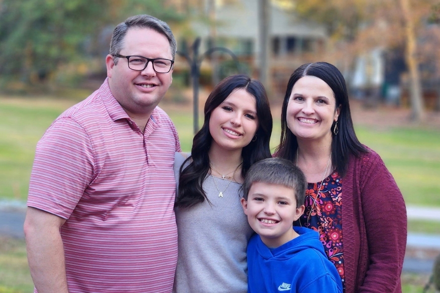 A family of four posing outdoors for a portrait. The parents stand on the outside, framing their two children—a teenage daughter and a younger son—in the center. All are smiling warmly.