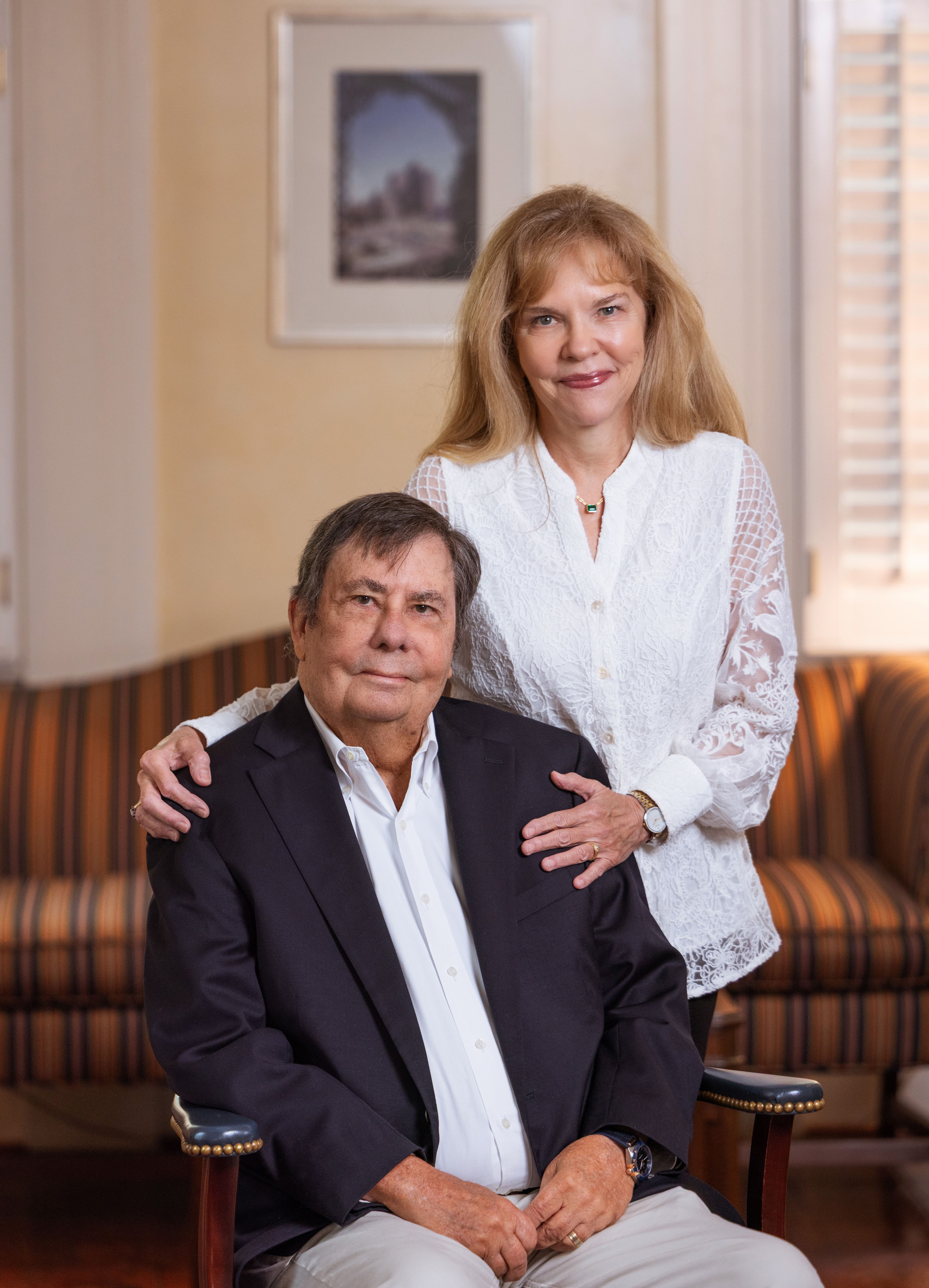 A man seated in formal wear and woman standing behind him in white blouse