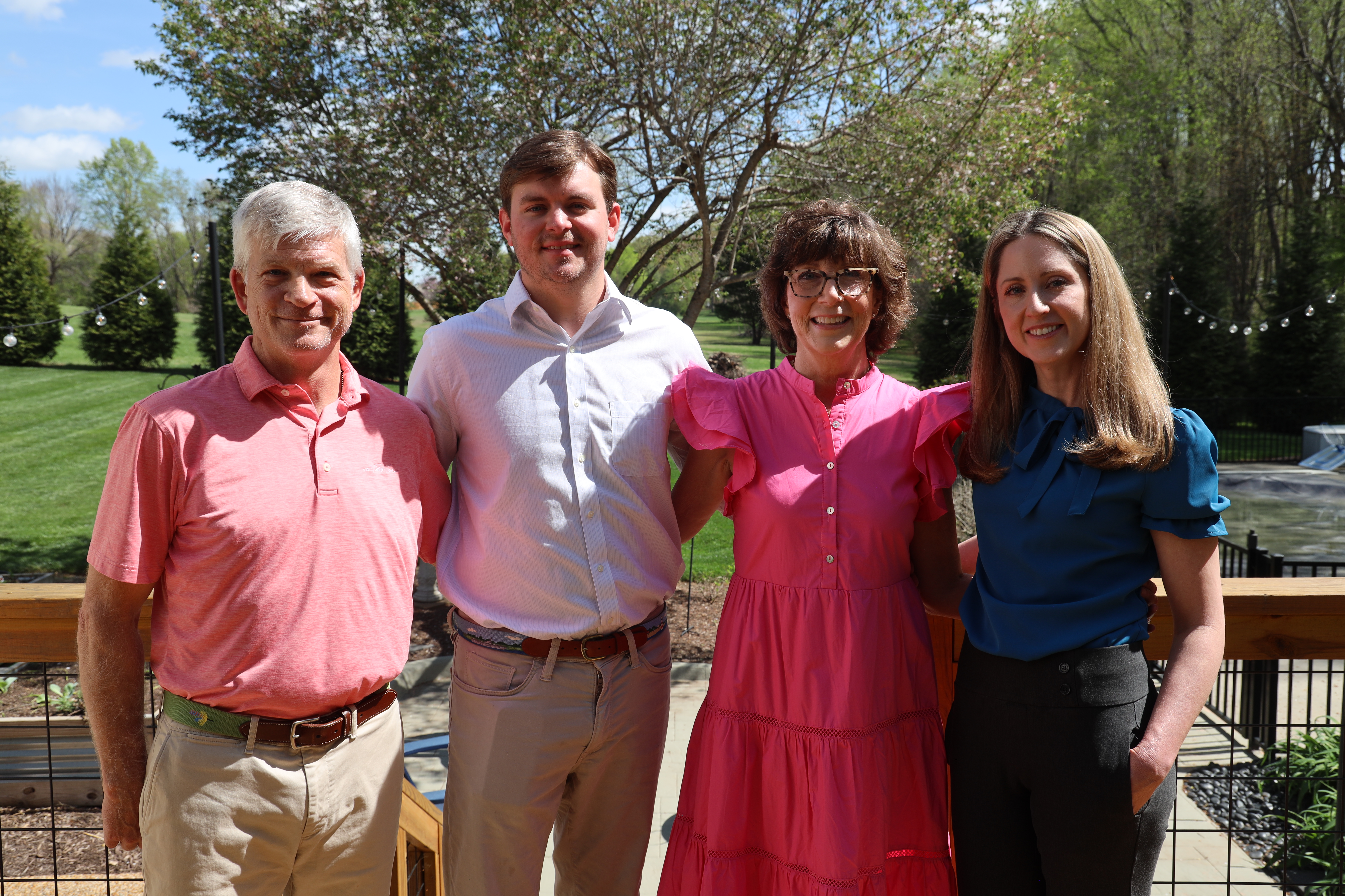 A group of four adults—two men and two women—standing together and smiling for a portrait on an outdoor wooden deck. The background shows a lush green lawn, trees, and a clear blue sky.