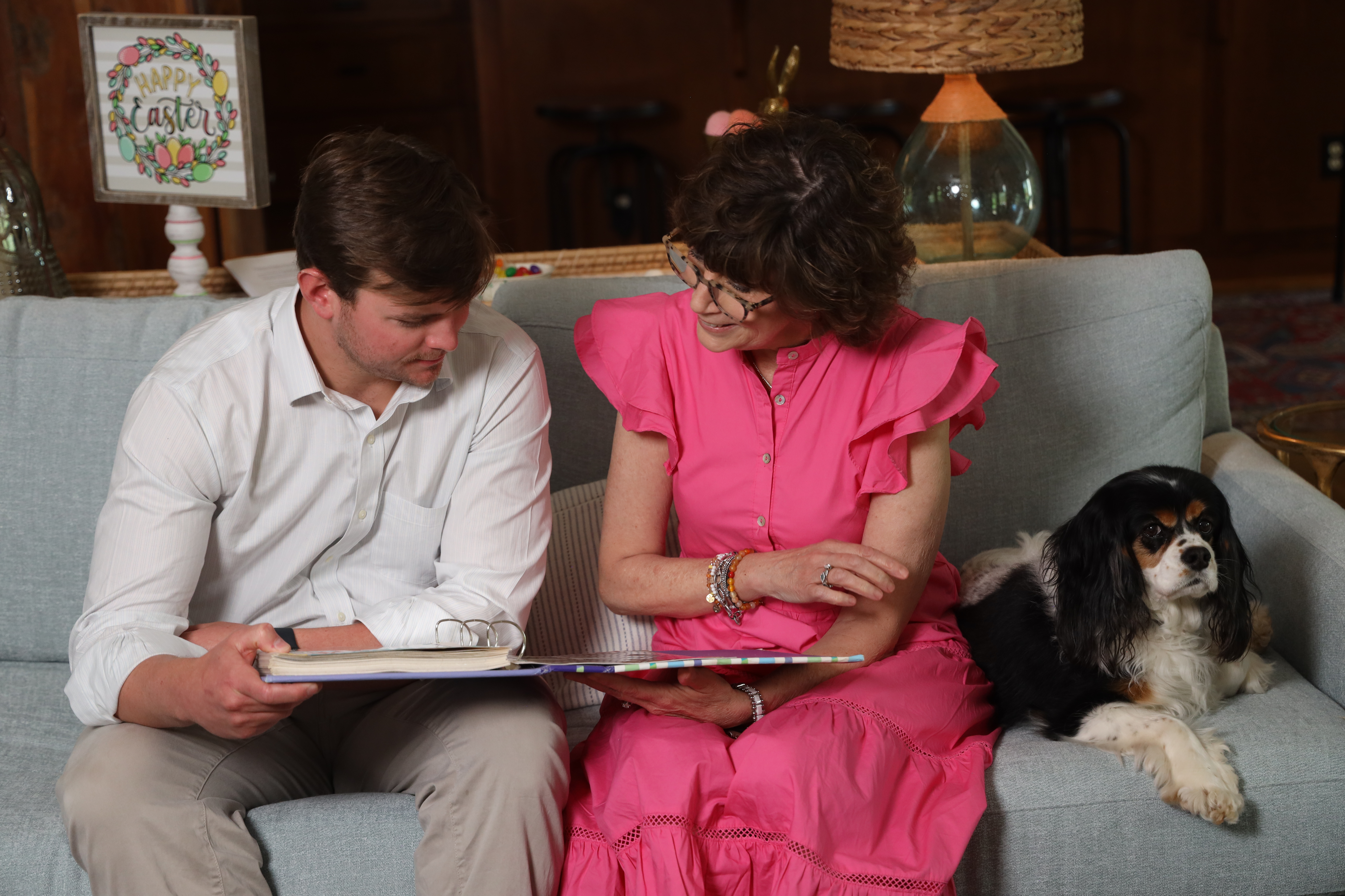 A man and a woman sitting on a light blue couch, looking through a large photo album together. A small black, white, and brown dog sits beside them, and a 