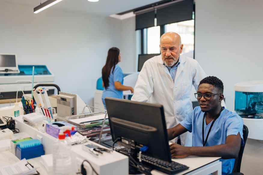 A senior doctor in a white coat leaning over to review computer screen results with a young male technician in blue scrubs in a modern medical laboratory.