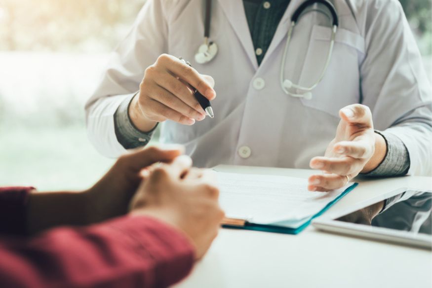 A close-up view of a doctor in a white lab coat and stethoscope gesturing with a pen while consulting with a patient sitting across a desk with medical documents and a tablet.