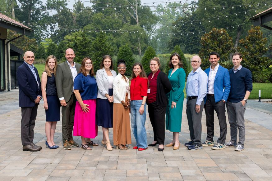 A group of eleven professionals standing in a line side-by-side outdoors on a paved patio, posing together in business-casual attire with trees and string lights in the background.