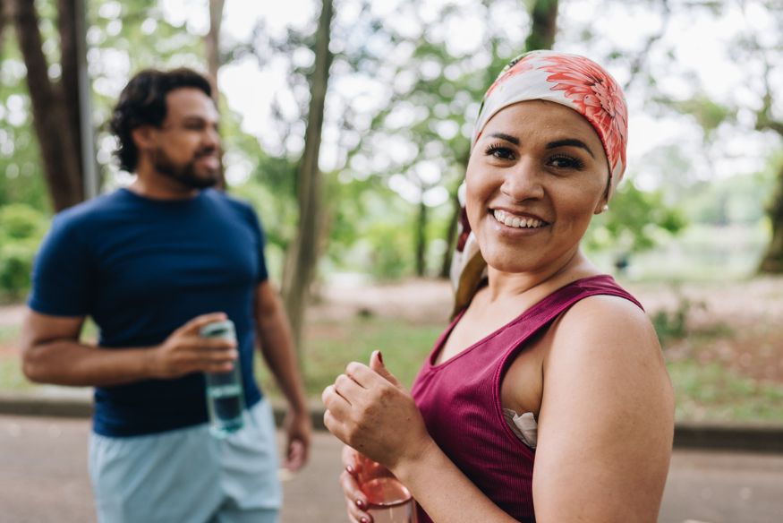 A smiling woman wearing a floral headscarf and tank top holds a water bottle during an outdoor workout, with a man standing blurred in the background