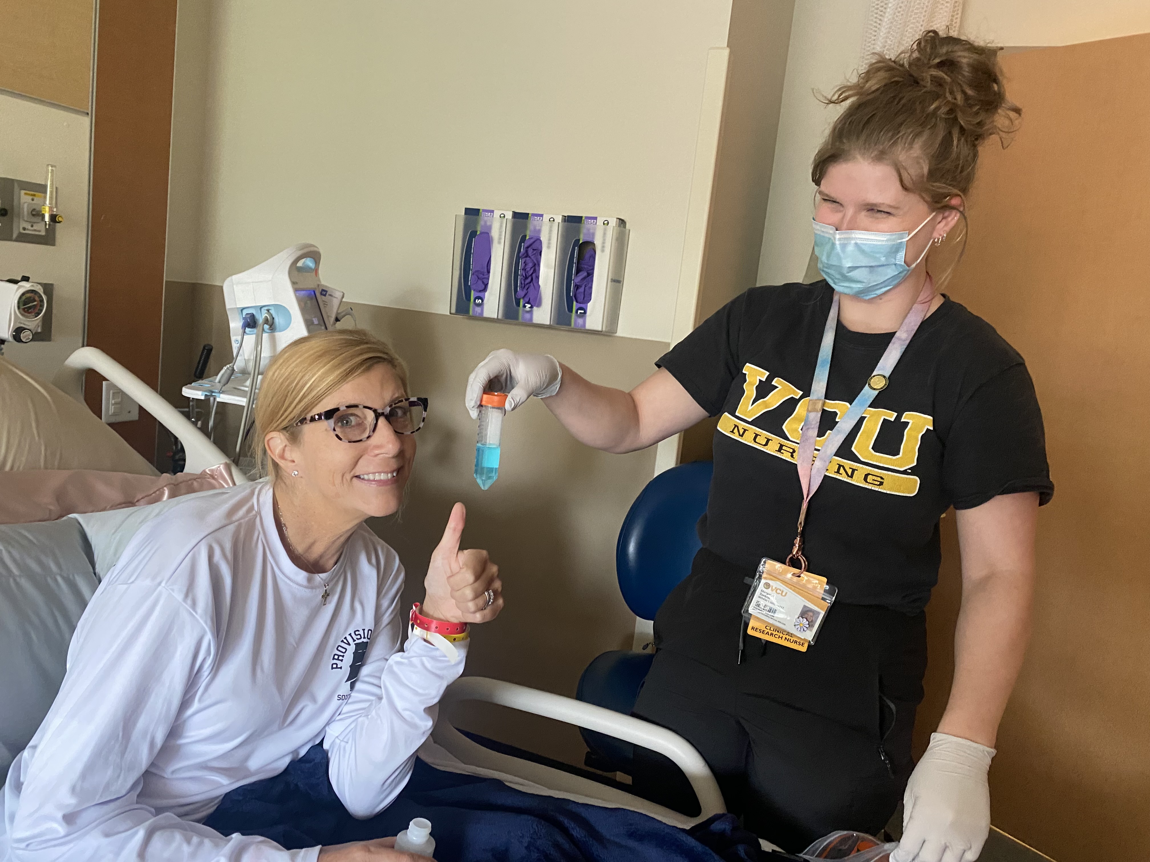 A woman in a hospital bed giving a thumbs-up while a nurse in a black VCU Nursing shirt holds up a small tube containing a bright blue liquid.