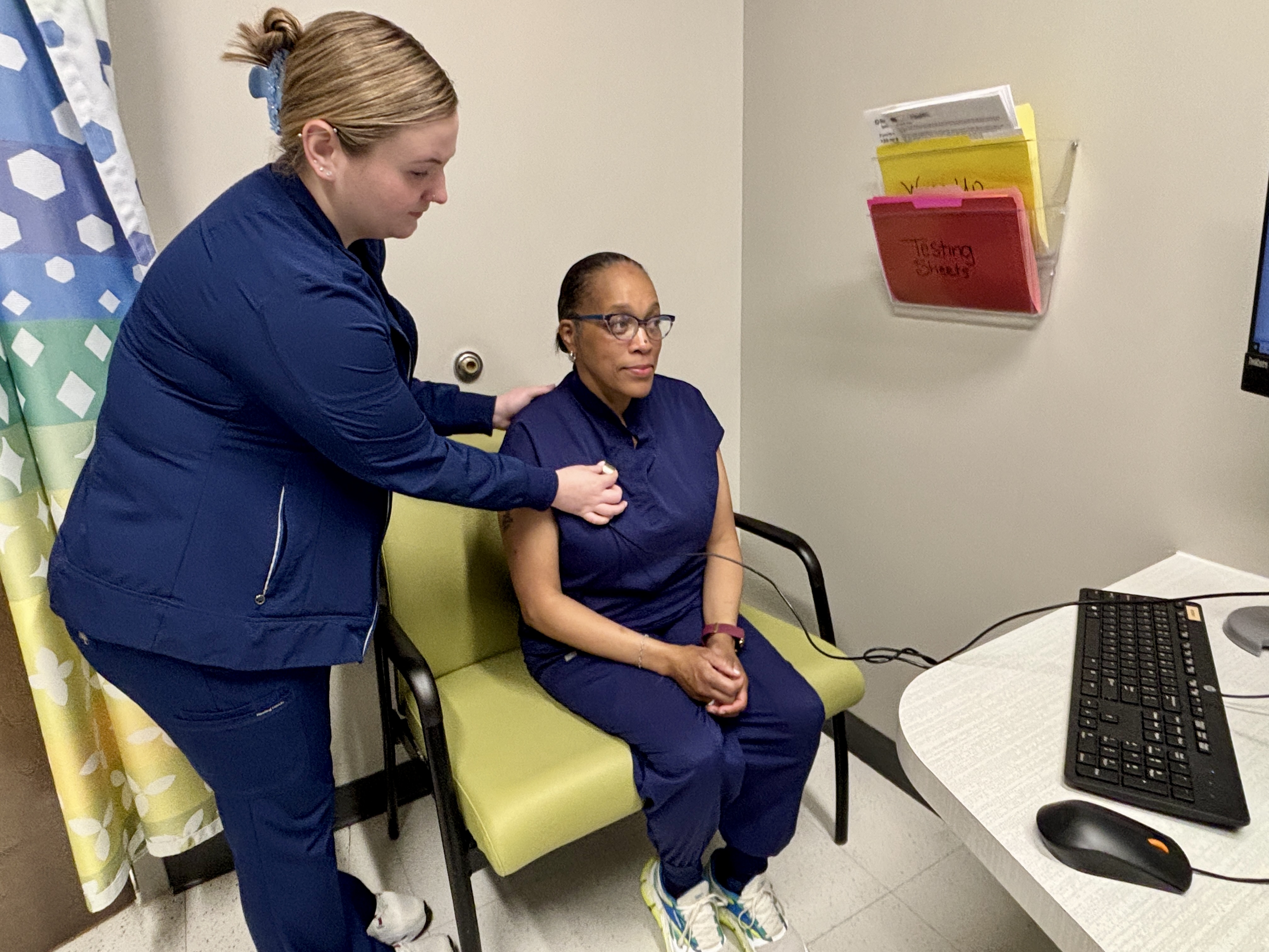A healthcare professional in blue scrubs uses a digital stethoscope to examine a patient seated in a chair within a medical office.