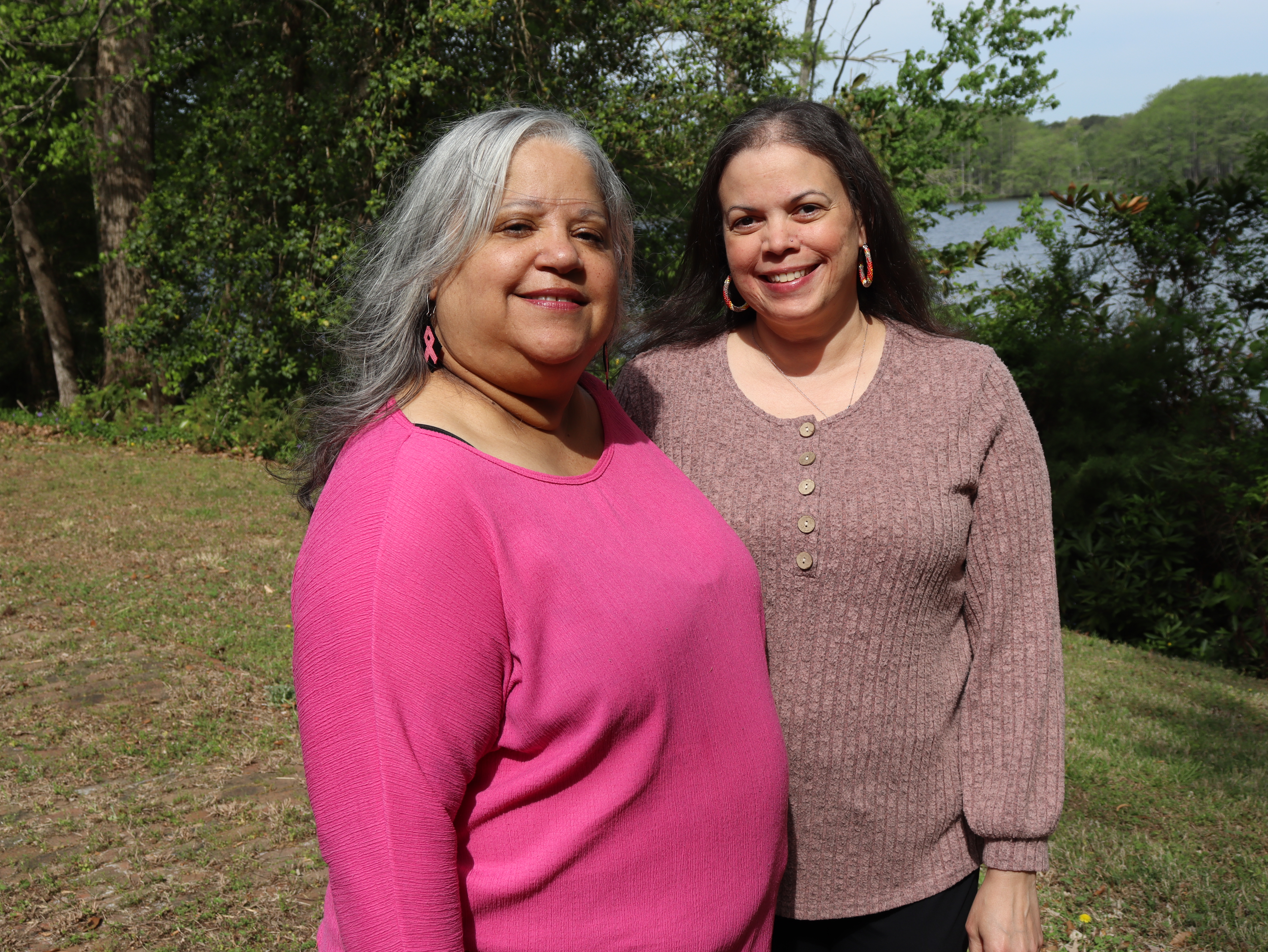 A medium shot of Melanie R. Stewart, wearing a bright pink shirt and pink breast cancer awareness ribbon earrings, standing outside alongside her daughter, Lindsey Johnson. Lindsey is wearing a mauve, textured long-sleeve shirt and smiling broadly. They are standing on a grassy area with green trees and water visible in the background.