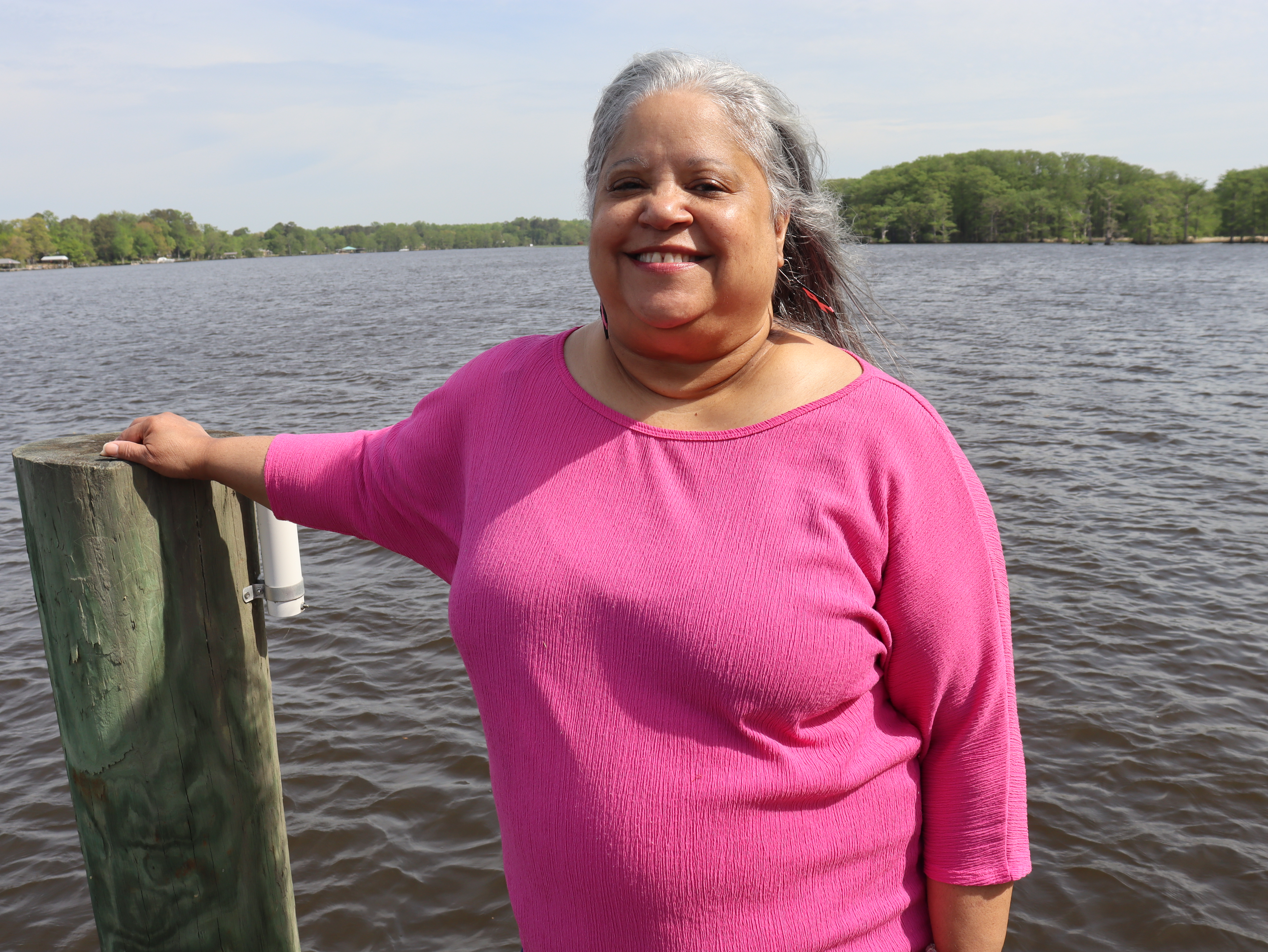 A medium shot of Melanie R. Stewart, a smiling woman with long, grey-streaked hair. She is wearing a bright pink long-sleeve shirt and standing outside next to a large body of water, resting her right hand on a wooden dock post. A green, tree-lined shoreline stretches across the background under a pale blue sky.