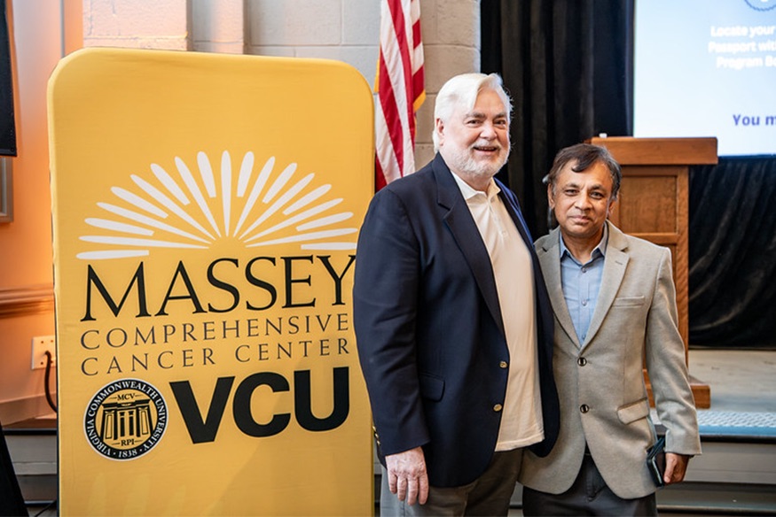 Two men posing together next to a yellow Massey Comprehensive Cancer Center VCU banner indoors.