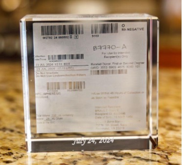 A clear, acrylic keepsake block encasing a medical label for a blood or marrow donation. The label includes barcodes, the blood type 