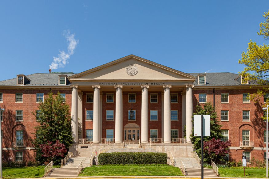 Exterior view of the National Institutes of Health (NIH) brick building featuring prominent white neoclassical columns and a large entrance staircase under a clear blue sky.