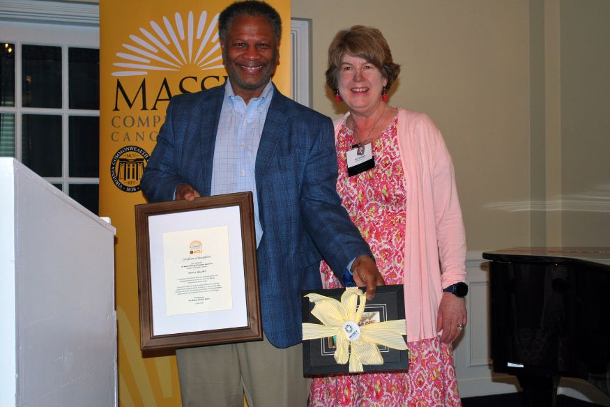 Robert Winn and Amy Williams smiling together indoors while holding a framed certificate and a gift wrapped with a yellow ribbon.