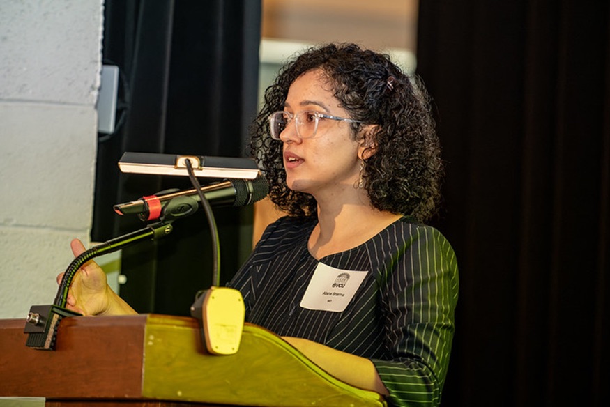 Alisha Sharma speaking into a microphone at a wooden podium during a presentation, wearing a black pinstriped top and glasses.