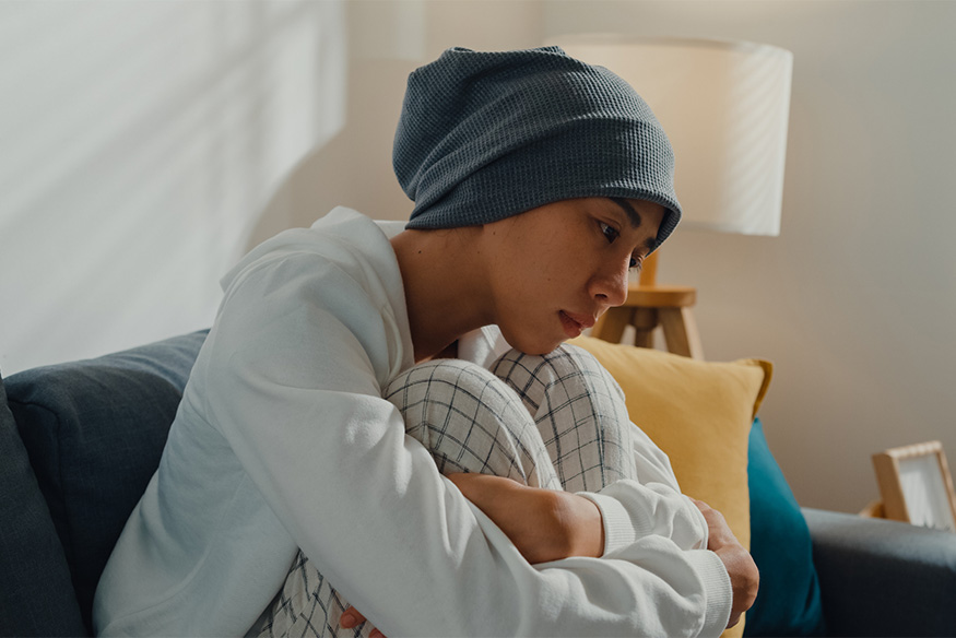 A young person wearing a grey beanie beanie and white hoodie sits on a couch, curled up with knees to chest and a thoughtful expression.