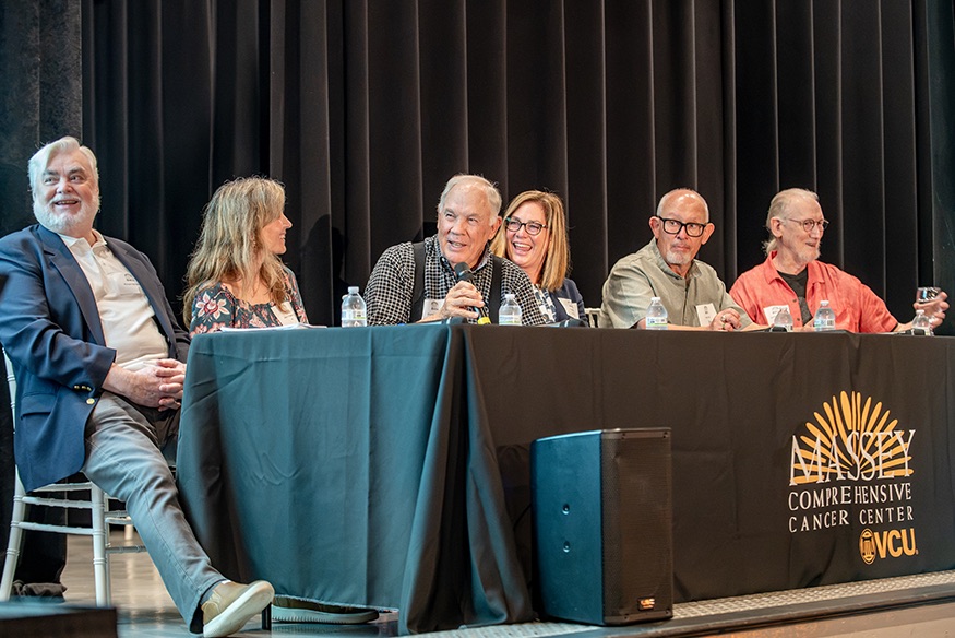 Six panelists seated at a long table with a black tablecloth branded with the Massey Comprehensive Cancer Center VCU logo during an event on stage.