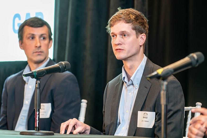 John Melson seated at a panel discussion table with a microphone in front of him, speaking alongside another attendee.