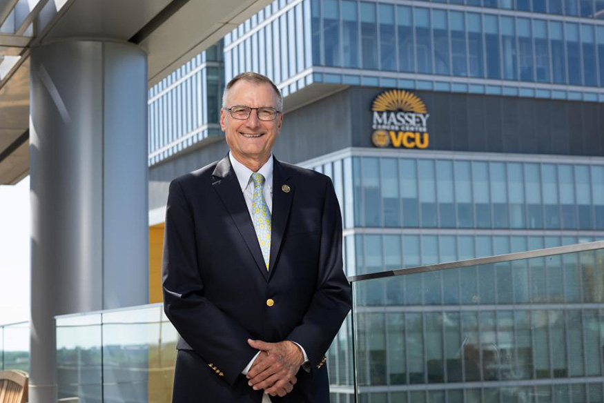 A smiling man in a dark suit jacket, white shirt, and patterned yellow tie stands outdoors on a balcony. In the background is a modern glass building featuring the 