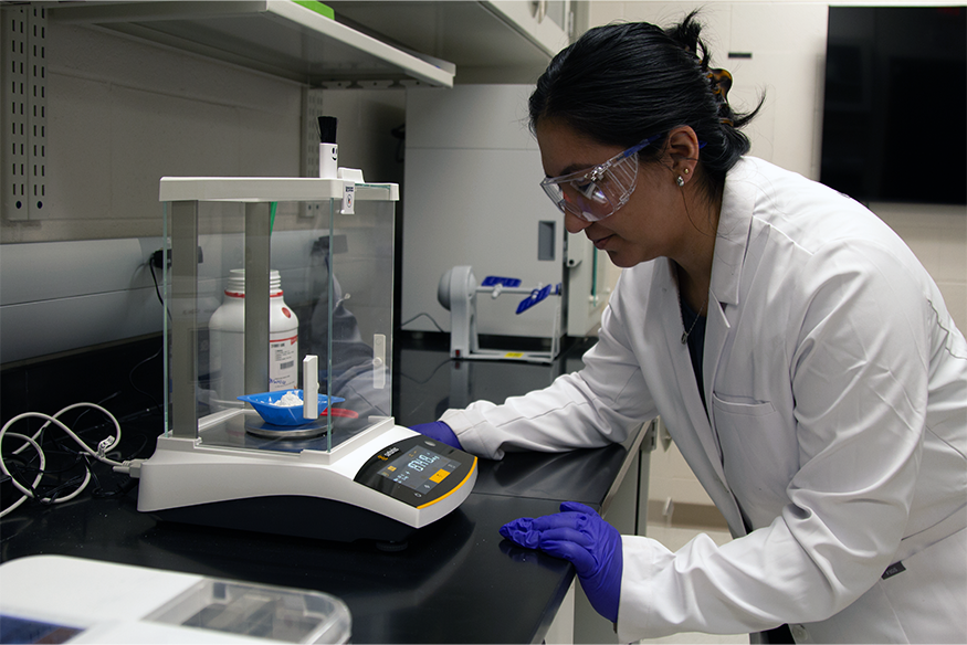 A scientist wearing a white lab coat, safety glasses, and purple gloves leans forward to read the digital display of an analytical balance scale while weighing a white powder sample in a laboratory.